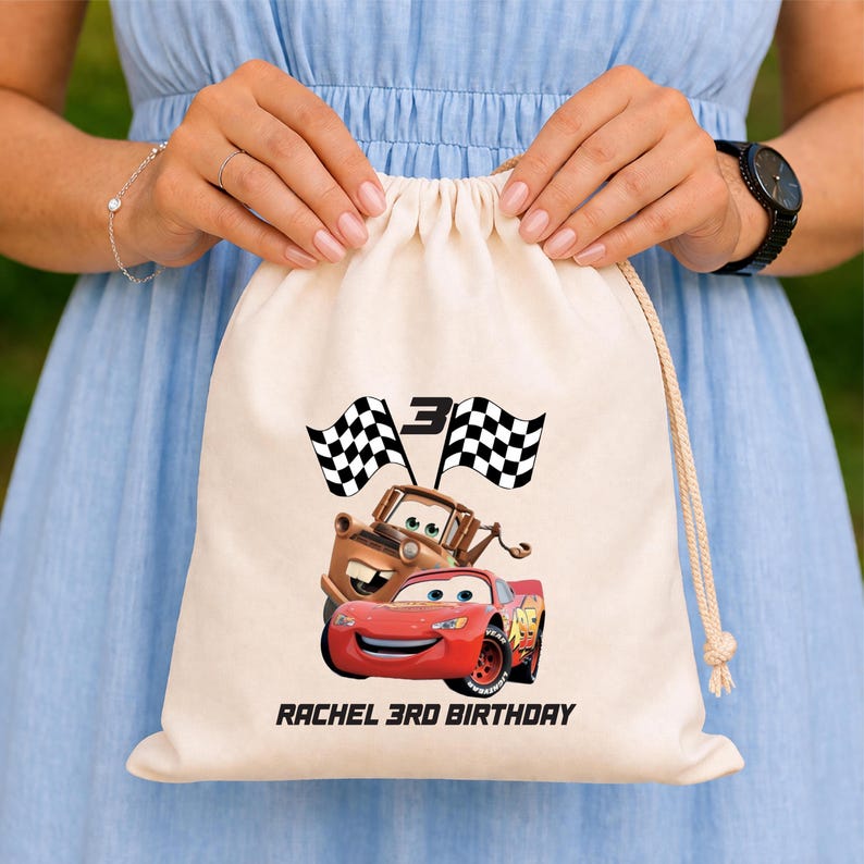 May include: Cream-coloured drawstring bag with a red race car and a brown truck, plus checkered flags and the number 3. The text "RACHEL 3RD BIRTHDAY" is printed below. The bag is held by a person.