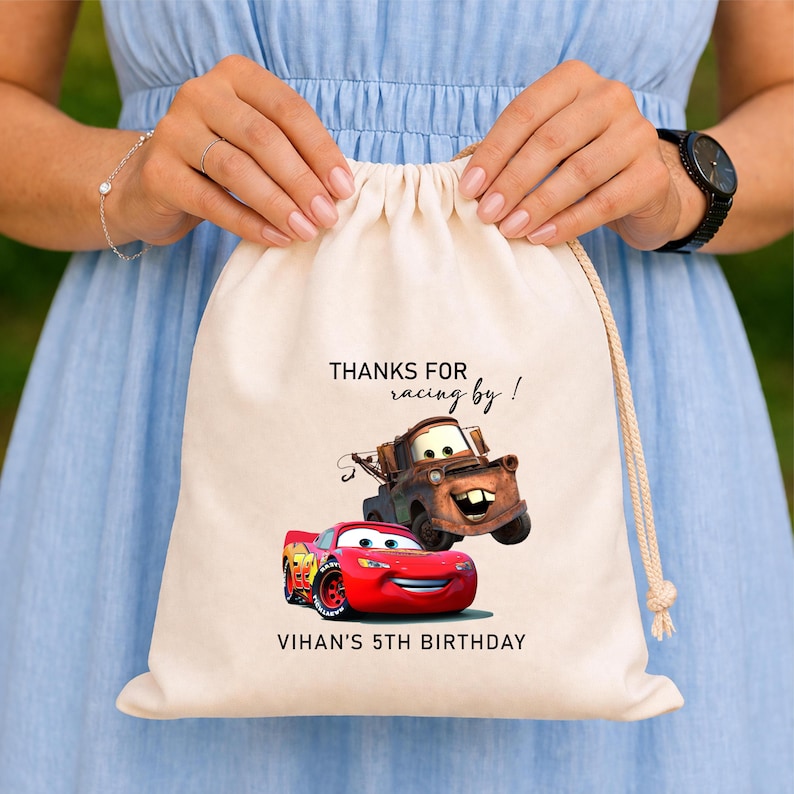 May include: A beige drawstring bag with the text "THANKS FOR racing by!" and "VIHAN'S 5TH BIRTHDAY". The bag displays a cartoon image of a red race car and a brown tow truck.
