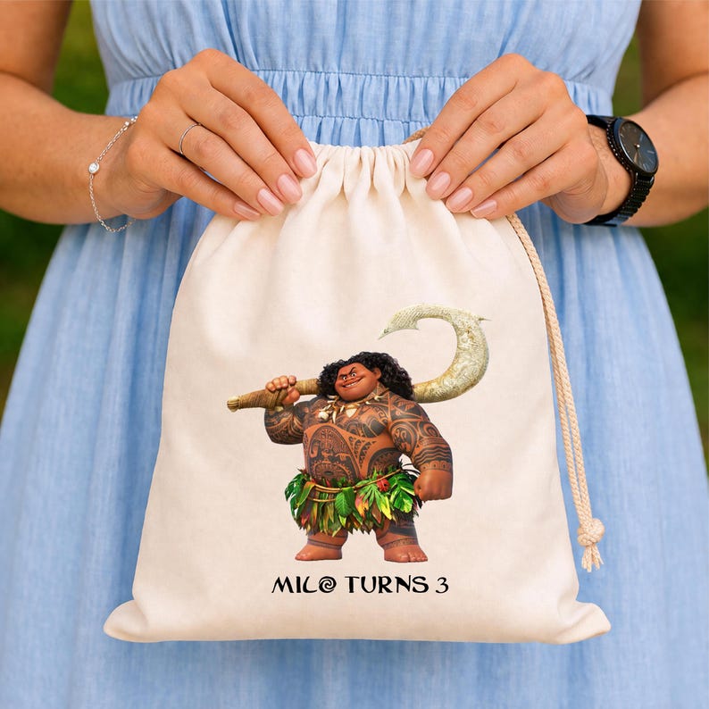 May include: A beige drawstring bag featuring a colorful illustration of the character Maui from the animated film Moana. Maui is depicted holding his magical fish hook. The text "MILO TURNS 3" is printed below the character.