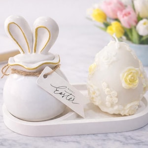 Może przedstawiać: Biały ceramiczny pojemnik w kształcie królika ze złotymi akcentami i etykietą z napisem "Happy Easter" stoi na białej tacy obok świecy w kształcie jajka ozdobionej żółtymi różami. W tle bukiet tulipanów.