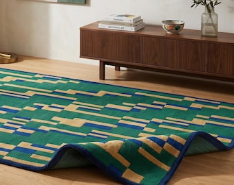 Puede incluir: Una alfombra rectangular con un patrón geométrico de bloques rectangulares verdes, beige y azules. La alfombra tiene un borde azul oscuro y una esquina está doblada para revelar la parte inferior, mostrando el patrón y la construcción.
