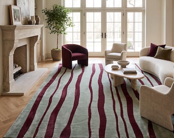 Puede incluir: Una alfombra rectangular grande con un fondo verde salvia claro y rayas verticales onduladas de color burdeos oscuro. La alfombra está colocada sobre un suelo de madera en espiga claro en una habitación con chimenea y muebles.
