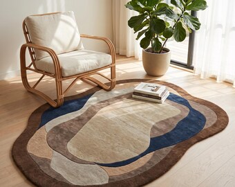 Puede incluir: Una alfombra de forma ovalada irregular con un patrón abstracto en capas en tonos marrón oscuro, azul marino, beige y gris claro. La alfombra tiene un pelo suave y texturizado y está colocada sobre un suelo de madera clara.