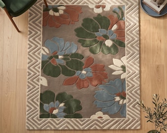 Puede incluir: Una alfombra rectangular con fondo color topo y grandes motivos florales con relieve en tonos verde bosque, terracota y azul empolvado. La alfombra está enmarcada por un borde ancho con patrón de chevrón en crema y topo con acabado texturizado.