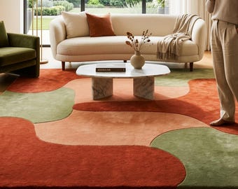 Puede incluir: Una alfombra grande con un patrón abstracto de formas orgánicas y fluidas en tonos terracota, naranja quemado, verde salvia y beige suave. La alfombra tiene un pelo texturizado y denso que cubre una parte importante del suelo de madera.