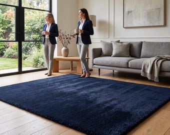 Puede incluir: Una alfombra grande y rectangular de color azul marino profundo con una textura gruesa, afelpada y de pelo largo. La alfombra está colocada sobre un suelo de madera clara, cubriendo gran parte del espacio frente a un sofá gris y un banco.