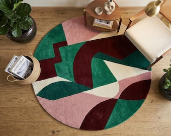 May include: A circular area rug featuring a bold abstract pattern with large, curved and geometric shapes in deep burgundy, forest green, soft pink, and cream. The rug has a textured, plush pile surface and is displayed on a light wood floor.
