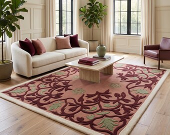 Puede incluir: Una alfombra rectangular con fondo rosa empolvado y un borde destacado de motivos florales en espiral de color burdeos oscuro y verde salvia. La alfombra tiene un borde grueso de color crema y un acabado de pelo suave y texturizado.