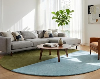 May include: A large gray sectional sofa with a chaise, a round wooden coffee table, and a tan leather armchair. A circular rug features a split design of olive green and light blue. A potted plant and abstract wall art complete the living room.