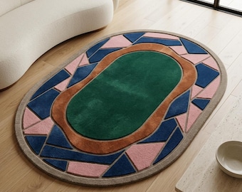Puede incluir: Una alfombra ovalada con un campo central verde oscuro rodeado por un borde grueso y ondulado de color marrón. El borde exterior presenta un patrón geométrico de formas azul oscuro y rosa claro, todo enmarcado por un borde beige sólido.