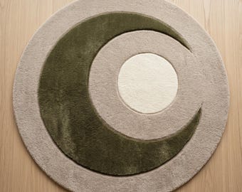 May include: A circular area rug featuring a tufted texture with a beige background. The design includes a large, dark olive green crescent moon shape that curves around a central, solid off-white circle, creating a layered celestial motif.
