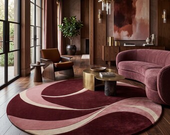 Puede incluir: Una alfombra circular grande con un patrón de ondas abstractas superpuestas en tonos burdeos intenso, rosa apagado y crema. La alfombra tiene una textura lisa de pelo corto y está colocada sobre un suelo de madera oscura.