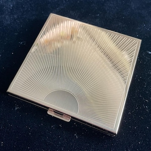 May include: A gold-toned, square compact featuring a sunburst pattern emanating from a semi-circular base. The compact is rectangular with a small clasp. The surface is reflective, and the design is etched into the metal, giving it a vintage look.