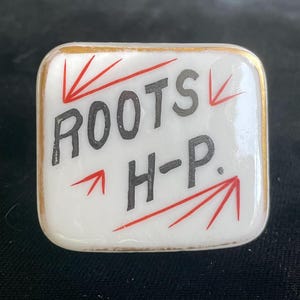 May include: A white, square ceramic tile with gold trim. The tile features the words "ROOTS H-P" in black lettering, with red arrows pointing towards the text. The design is simple and the tile has a glossy finish.