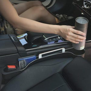 May include: A black car seat organizer with a cup holder, a slot for a phone, and a small pocket. A person is holding a gray insulated tumbler. A blue wallet and car keys are also visible.