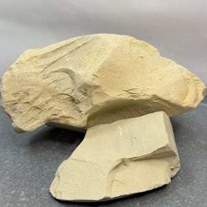 May include: Two pieces of raw, beige clay. The larger piece is irregularly shaped with a rough texture, while the smaller piece is rectangular. The clay appears to be natural and unprocessed, with visible variations in colour and surface.