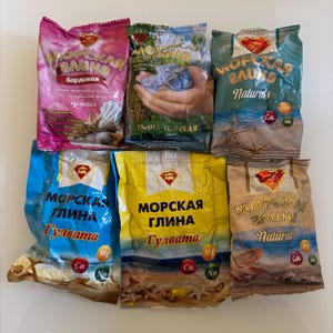 Puede incluir: Una variedad de paquetes coloridos de productos de arcilla marina. Los paquetes son de varios colores, incluyendo rosa, verde, azul y amarillo, con texto cirílico e imágenes del mar. Contiene minerales como Ca, Mg y Na.