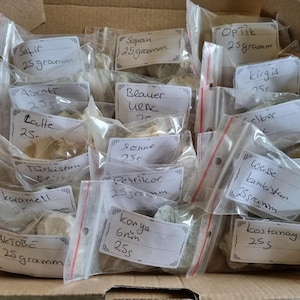 May include: A collection of small, clear plastic resealable bags filled with various mineral or clay samples, each labelled with handwritten text indicating names like Safir, Sapan, OpTik, Kirgis, Asrotr, Blauer Urac, Gelber, Latte, Sonne, Turkistan, Petrikor, Weisse, Karamell, Konya, Aktobe, and Kostanay, with most bags marked as 25 grams.