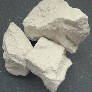Peut inclure: Trois morceaux de craie blanc cassé de forme irrégulière sont disposés sur une surface gris foncé. Les morceaux de craie ont un aspect rugueux et texturé, avec des arêtes et des crevasses visibles. L'éclairage met en valeur les variations naturelles de la surface de la craie.
