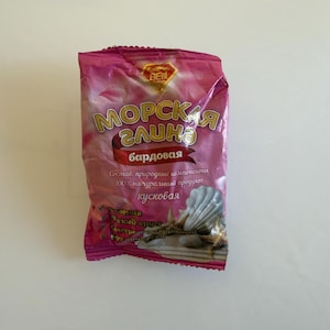 May include: A pink package of "Sea Clay" with Russian text. The package features gold lettering and a red banner with more text. The bottom of the package has a seashell and starfish design.