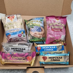 Op de afbeelding: Een kartonnen doos met zes verpakkingen kleiproducten. De items bevatten drie zakjes met het label MOPCKAЯ ГЛИHA in beige, groen en roze, en drie rechthoekige repen met het label MOPCKAЯ ГЛИHA of БATOНЧИK БEHTOHИTOBЫЙ in wit, blauw en paars.