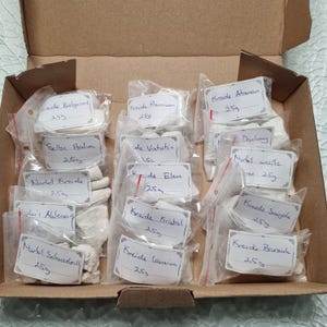 May include: A cardboard box containing fourteen small, clear plastic bags filled with white chalk chunks. Each bag features a white label with handwritten text identifying the variety and a weight of 25g, including names like Belgorod, Premium, Ataman, and others.