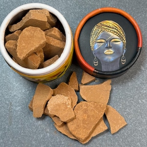 Könnte beinhalten: Offener Behälter mit hellbraunen, unregelmäßig geformten Stücken, möglicherweise ein Kosmetikprodukt. Der Behälter hat ein gelbes und orangefarbenes Design. Ein separater Deckel zeigt ein gemaltes Gesicht mit goldenen Akzenten und Schmuck.