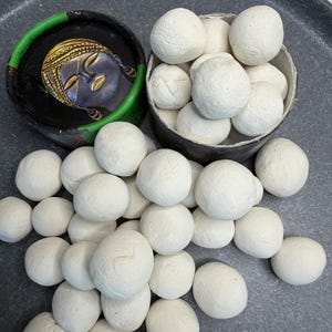 Op de afbeelding: Een verzameling matte witte, bolvormige kleiballen verspreid rond een cilindrische houder gevuld met meer kleiballen. Naast de houder staat een klein, rond zwart doosje met een goudkleurige illustratie van een sereen gezicht.