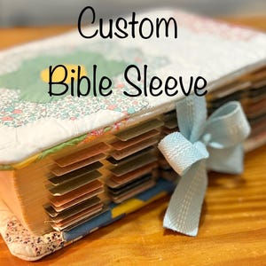 May include: A quilted fabric Bible sleeve featuring a floral pattern with a light blue ribbon bow closure. The sleeve contains a book with numerous colored page tabs visible along the side. The text Custom Bible Sleeve is printed on the image.