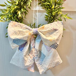 May include: A decorative bow crafted from patchwork fabric featuring a variety of light blue, white, and soft orange patterns. The bow is constructed with quilted squares and is suspended by a thin white string, showcasing a textured, multi-patterned surface.