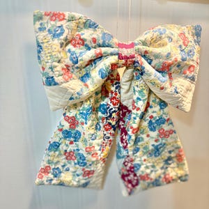 May include: A large decorative bow crafted from quilted fabric featuring a multi-colored floral pattern with red, blue, yellow, and green blossoms on a cream background. The center knot is a solid pink fabric, and the bow hangs from a thin string.