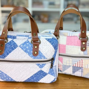 May include: Two rectangular quilted handbags featuring brown leather handles secured with brass rivets. One bag displays a blue and white geometric pattern, while the other features a multi-colored pastel patchwork design. Both include a front zipper.