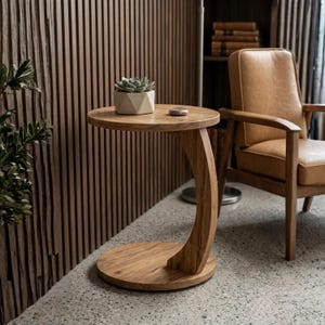 May include: A small, round wooden side table with a curved leg and a circular base. A small succulent plant in a geometric pot and a small, round stone sit on the tabletop. The table is made of light-colored wood.