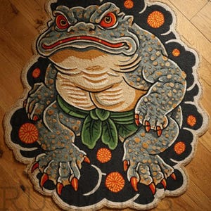 May include: A detailed rug featuring a stylized frog design. The frog is primarily gray with orange spots, red eyes, and a green sash. The rug has a black border with orange floral accents and is placed on a wooden floor.