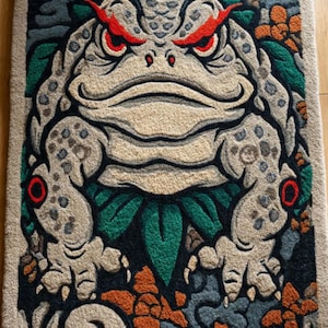 May include: A rectangular rug featuring a detailed illustration of a large, stylized frog. The frog is primarily beige with red eyes and accents of green, orange, and blue. The design has a textured appearance, suggesting a woven or tufted construction.