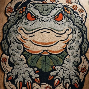 May include: A decorative rug featuring a detailed illustration of a frog with orange eyes and claws. The rug has a gray, green, and beige color scheme with orange flower accents. The frog design is set against a black background.