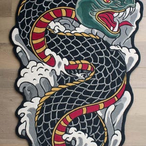 Op de afbeelding: Decoratief tapijt met een gestileerd drakenontwerp. De draak is zwart met rode, gele en groene accenten, tegen een achtergrond van grijze en witte golven. Het tapijt is gevormd om de vorm van de draak te volgen.