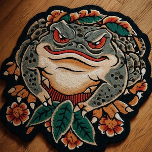 May include: A rug featuring a detailed illustration of a frog's face with red eyes, surrounded by floral and leaf accents. The rug has a black border and is placed on a wooden floor. The frog's face is primarily gray, with tan and orange details.