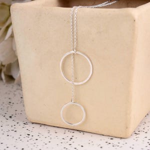 Puede incluir: Un collar plateado con dos círculos abiertos, uno encima del otro, conectados por una fina cadena. El collar se muestra sobre un fondo neutro, resaltando su diseño minimalista. Los círculos son de diferentes tamaños, creando un aspecto moderno y elegante.