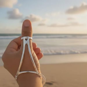 Può includere: Un set di anello e bracciale per pollice in argento. L'anello è collegato al bracciale da un design triangolare. I gioielli sono esposti su uno sfondo di spiaggia con l'oceano e il cielo sullo sfondo.