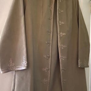 May include: An olive green coat with a mandarin collar and long sleeves. The coat has detailed embroidery along the front edges, cuffs, and collar. The fabric has a subtle ribbed texture. The coat is hanging on a hanger.