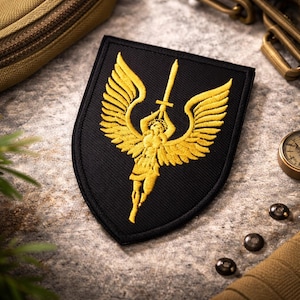 Peut inclure: Écusson brodé noir avec un ange doré tenant une épée et des ailes déployées. Le patch est en forme de bouclier. Le design est détaillé et les couleurs sont vives. Il est probablement destiné à une veste ou un sac.
