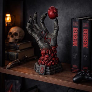 Puede incluir: Estatua detallada de una mano esquelética sosteniendo un orbe rojo. La mano es de color gris oscuro con detalles rojos y un símbolo en la palma. La base está adornada con pequeñas calaveras rojas. Dos libros "Berserk" están en el fondo.
