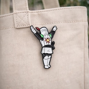 Puede incluir: Un pin de esmalte en blanco y negro de un caballero con los brazos levantados en una pose alegre. El caballero tiene una pluma roja, detalles verdes y un emblema de sol en el pecho. Una espada pequeña está unida al costado del caballero. El pin está en una bolsa de lona beige.