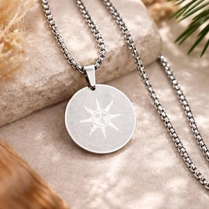May include: A silver-colored pendant necklace featuring a circular pendant with a sun design. The pendant hangs from a chain with a textured, rounded link pattern. The sun design has a face in the center with rays extending outward.