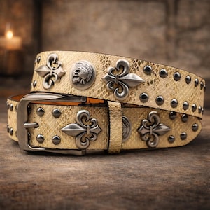 Pode incluir: Um cinto de couro bege com padrão de pele de cobra, adornado com flores de lis prateadas, medalhões de caveira e tachas. O cinto apresenta uma fivela prateada e foi concebido para um visual elegante.