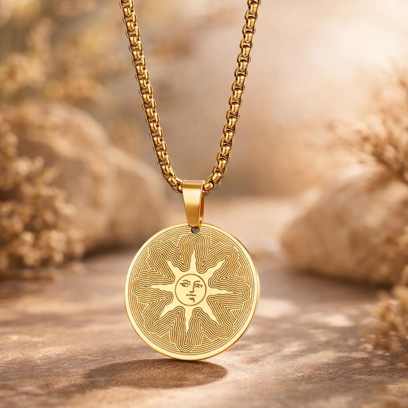 May include: Gold-colored pendant necklace featuring a circular pendant with a sun design. The sun has a face and radiating lines. The chain and pendant are a matching gold color, creating a cohesive and elegant look. The necklace is displayed against a neutral background.