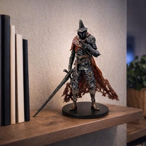 Op de afbeelding: Gedetailleerd beeldje van een krijger in donker pantser, met een groot zwaard. De figuur heeft een lange, puntige hoed en een gerafelde rode cape. Het staat op een zwarte, cirkelvormige basis, geplaatst op een houten plank. De figuur is ongeveer 25 cm hoog.