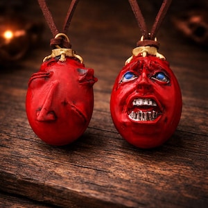 May include: Two red pendant necklaces with a face design. The pendants are oval-shaped with detailed facial features, including open mouths and blue eyes. They hang from brown leather cords, set against a wooden background.