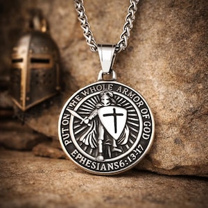 May include: A silver-colored pendant necklace featuring a circular medallion with a knight holding a sword and shield with a cross. The text "PUT ON THE WHOLE ARMOR OF GOD EPHESIANS 6:13-17" is inscribed around the edge. A chain and helmet are also visible.